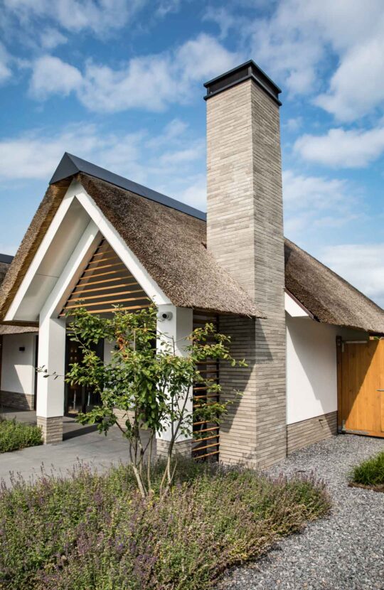 Landelijk-moderne woning met meerdere rieten kappen, houten gevelaccenten, wit stucwerk en een zorgvuldig aangelegde voortuin
