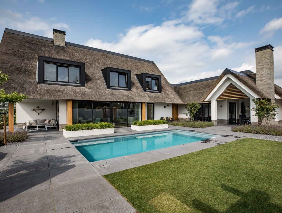 Tuinzijde van een villa met zwembad, grote glaspartijen, houten accenten en een rieten dak
