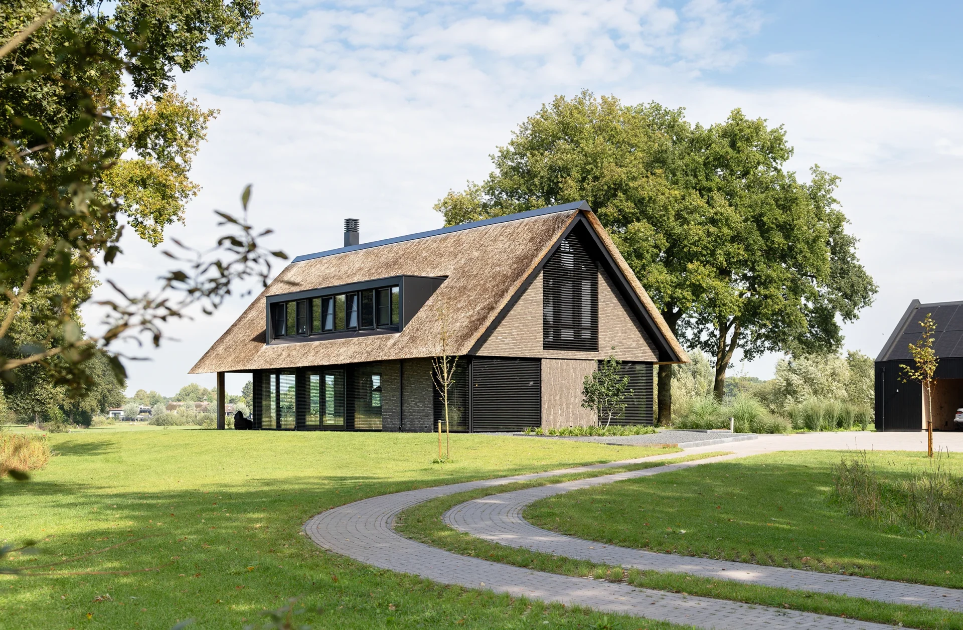 Moderne schuurwoning met rieten kap, grijze gevelstenen, donkere lamellen, houten accenten en grote raampartijen, ontworpen door Bekhuis & KleinJan.