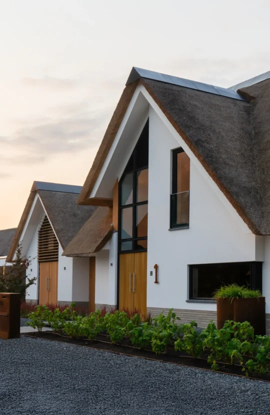 Entree van een landelijk-moderne villa met hoge houten voordeur, glaspartij, wit stucwerk en een rieten kap, ontworpen door Bekhuis & KleinJan