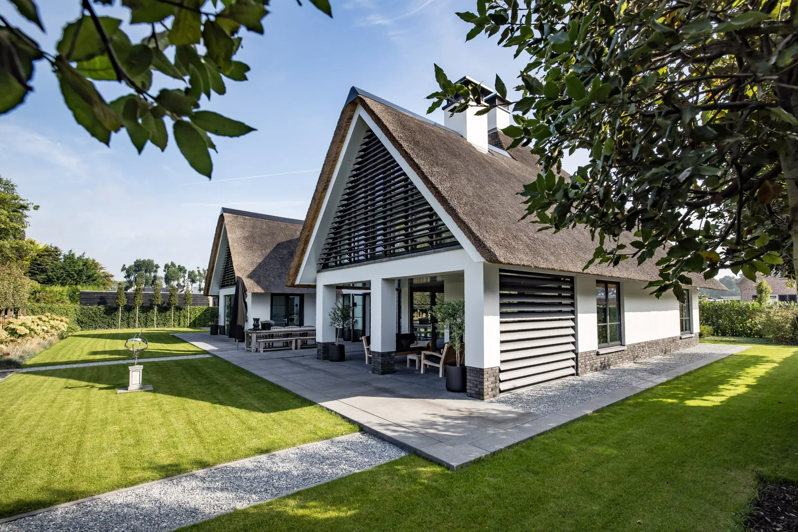 Villa met rieten dak en zwarte lamellen, gezien vanuit de groene tuin met bomen en een overdekt terras.