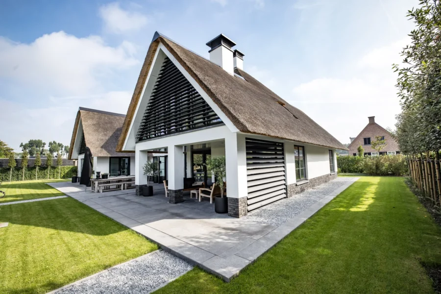 Zijaanzicht van een witte villa met rieten dak, zwarte lamellen, een overdekt terras en een strak aangelegde tuin.