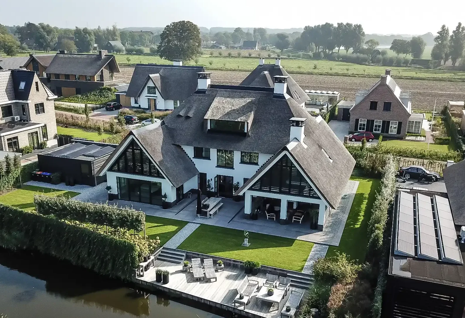 Royale villa met rieten dak, twee zijvleugels, een groene tuin en een houten terras aan het water.